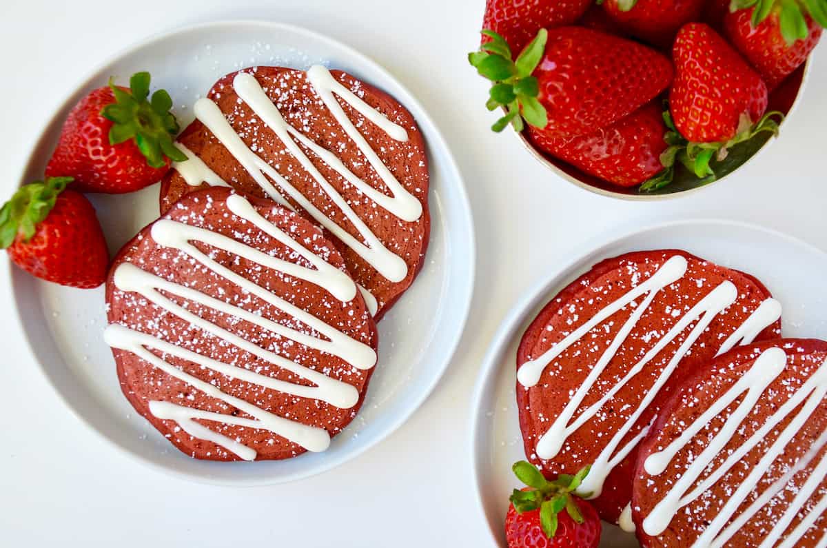 Red velvet pancakes drizzled with cream cheese glaze and sprinkled with powdered sugar are on white plates and garnished with strawberries. A bowl of strawberries sits nearby.