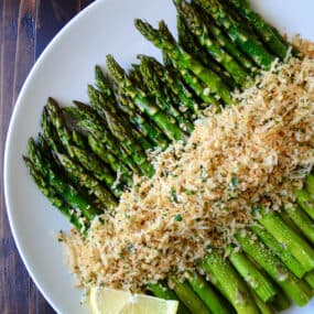 Roasted asparagus topped with cheesy, herby breadcrumbs on a white serving plate.