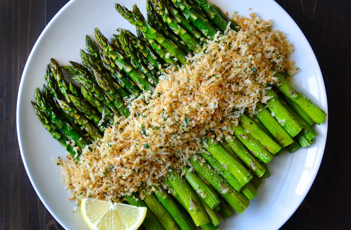 Roasted asparagus topped with cheesy, herby toasted breadcrumbs on a white, round serving plate.