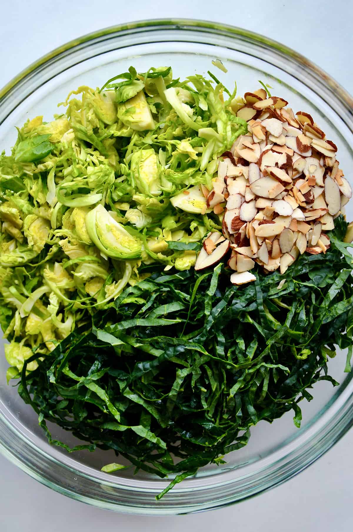 Shredded kale and Brussels sprouts and sliced almonds in a large glass mixing bowl.
