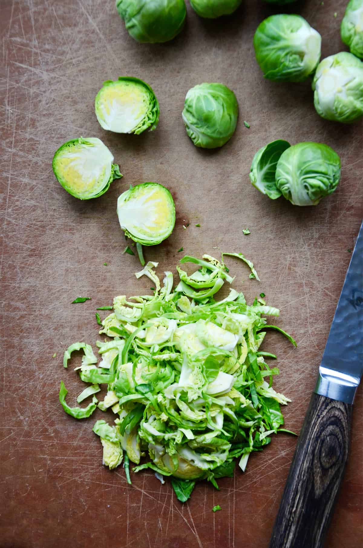Shredded and halved Brussels sprouts on a cutting board. A paring knife is to the side of the Brussels sprouts.