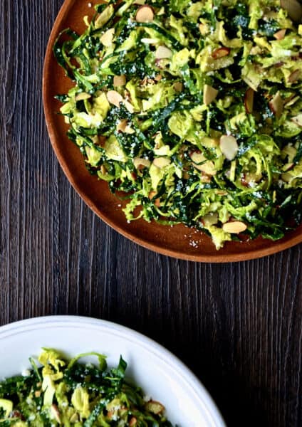 Two plates of shredded kale and Brussels sprouts salad with sliced almonds, topped with grated Parmesan cheese.