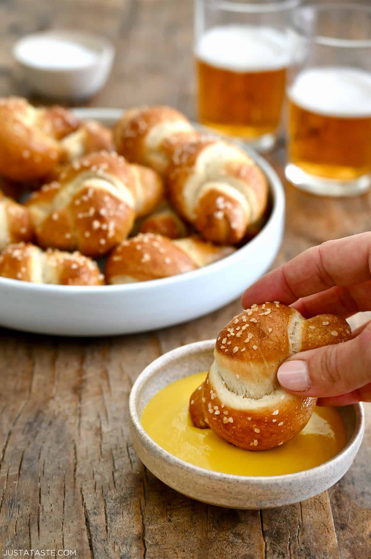 A hand holding a pizza dough soft pretzel knot dips it into cheese sauce. A bowl with soft pretzel knots is in the background.