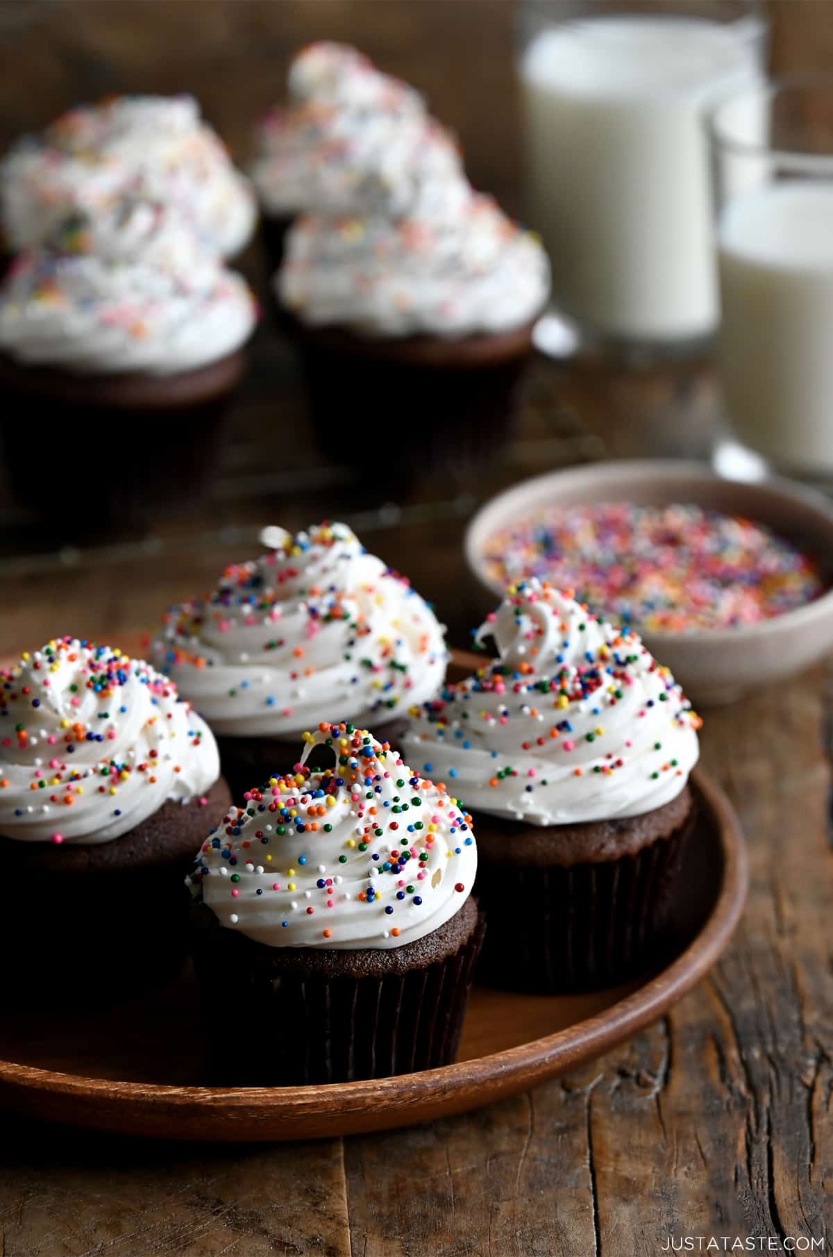 Four chocolate cupcakes topped with white frosting and rainbow sprinkles on a plate with more cupcakes on a wire rack in the background.