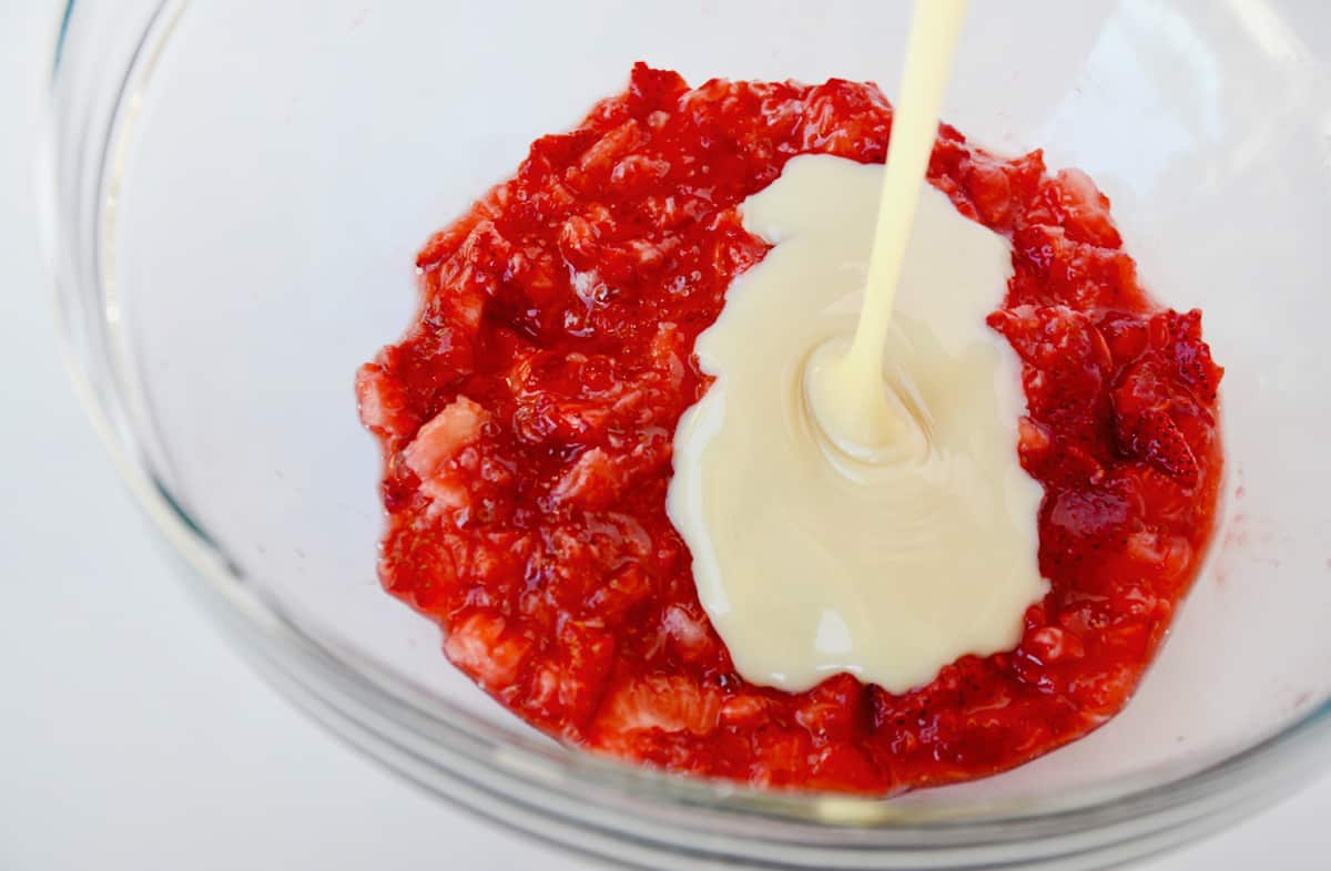 Sweetened condensed milk being added to mashed strawberries in a large glass mixing bowl.