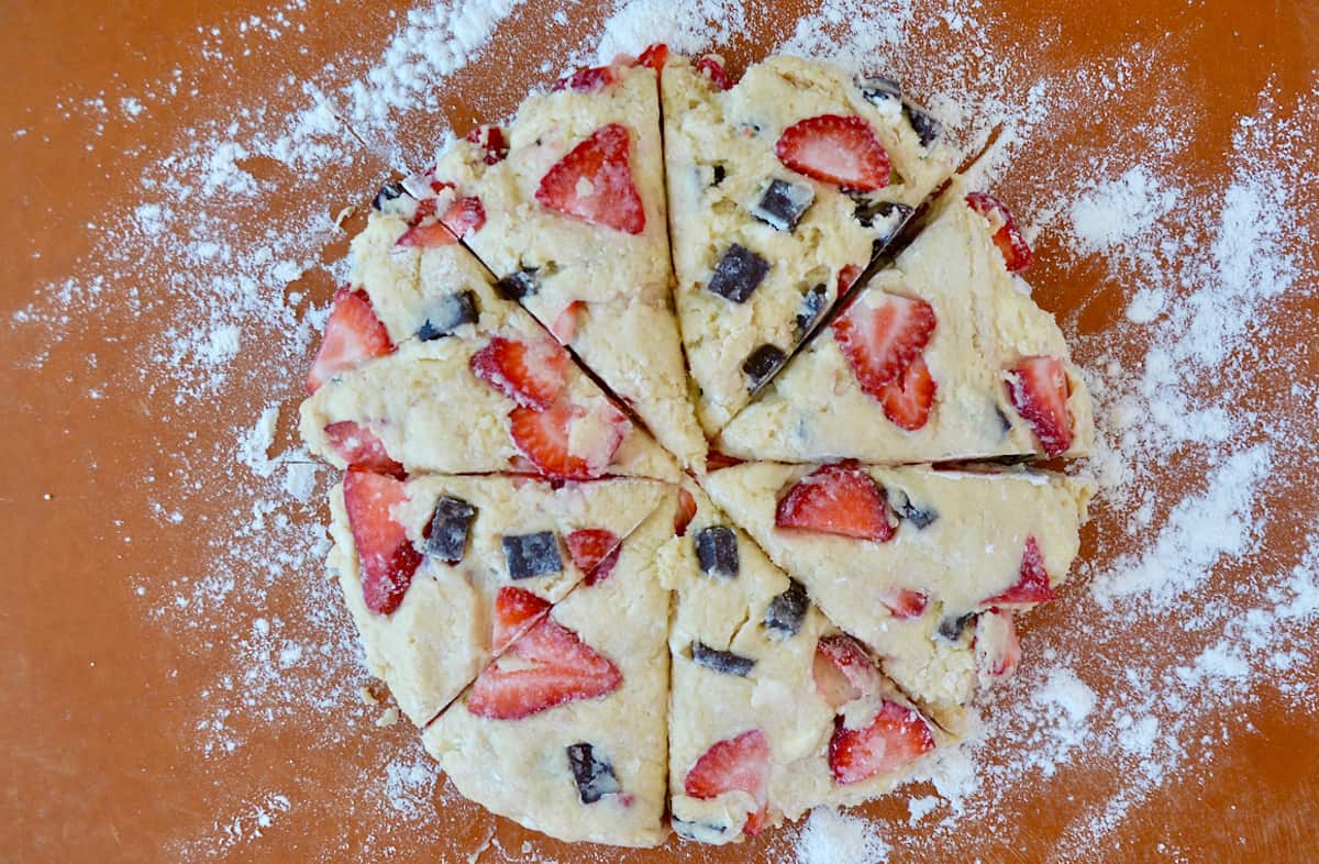 A disc of dough studded with strawberries and chocolate chunks sliced into eight wedges on a cutting board that's sprinkled with flour.