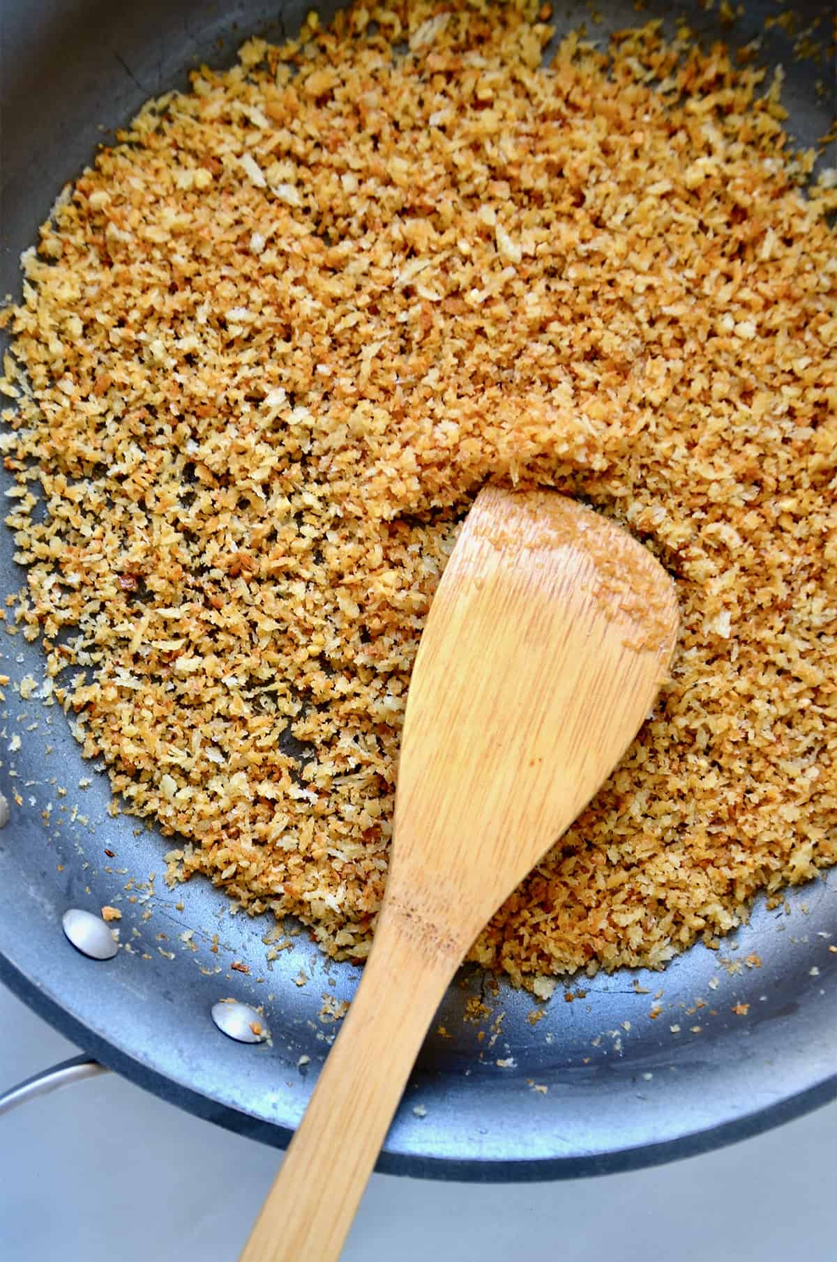 Toasted breadcrumbs in a nonstick skillet. A spoon rests against the side of the skillet.