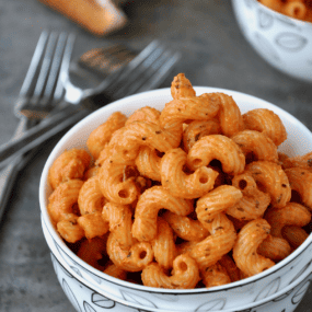 A small bowl of cavatappi pasta with pancetta and vodka sauce sits on a grey counter, with forks and crusty hunks of bread nearby.