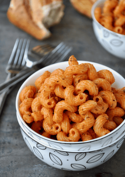 A small bowl of cavatappi pasta with pancetta and vodka sauce sits on a grey counter, with forks and crusty hunks of bread nearby.