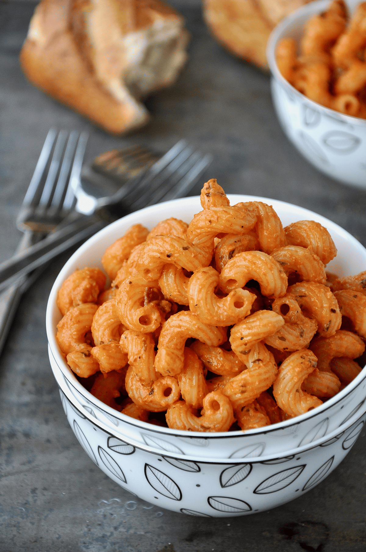 A small bowl of cavatappi pasta with pancetta and vodka sauce sits on a grey counter, with forks and crusty hunks of bread nearby.