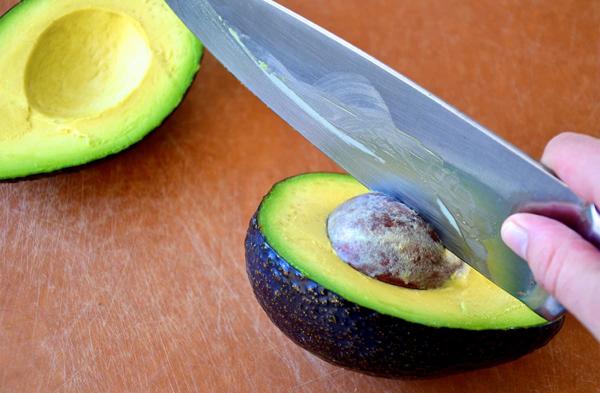 A person using a chef's knife to remove a pit from an avocado half.