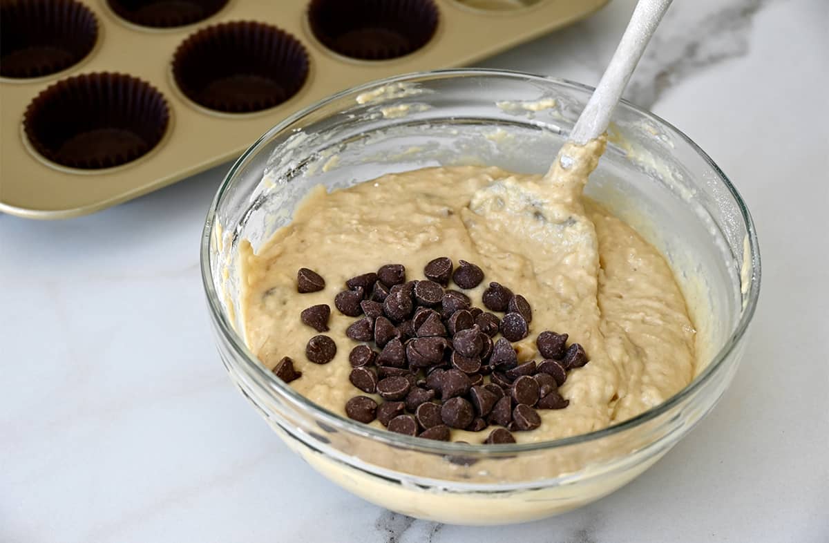 Banana muffin batter topped with chocolate chips in a glass bowl with a spatula next to a muffin tin with cupcake liners.