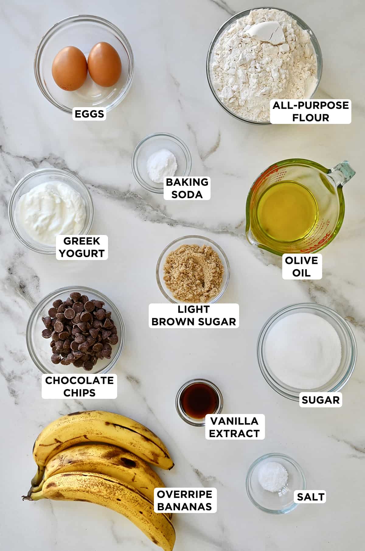 Overripe bananas next to various sizes of glass bowls and a liquid measuring cup containing the ingredients needed to make banana bread muffins, including flour, olive oil, sugar, vanilla extract, salt, chocolate chips, brown sugar, Greek yogurt, baking soda and eggs.