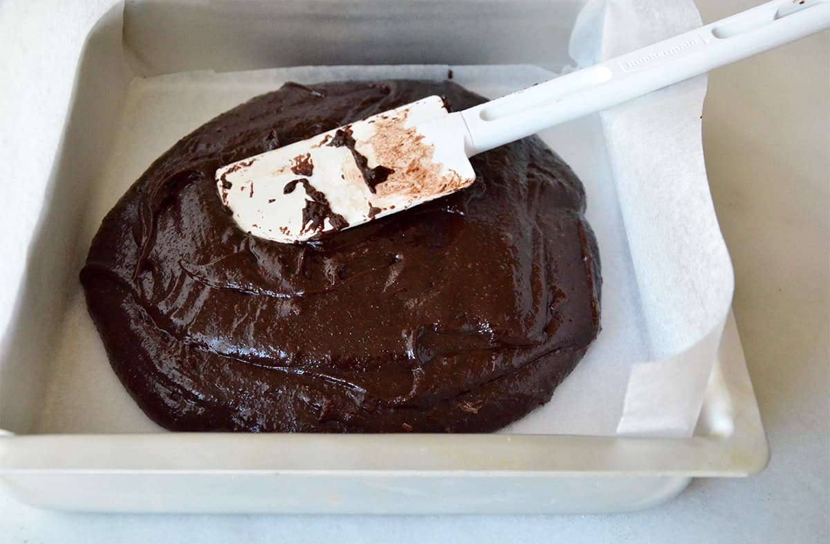 Brownie batter in a square baking dish lined with parchment paper. A rubber spatula rests against the side of the dish.