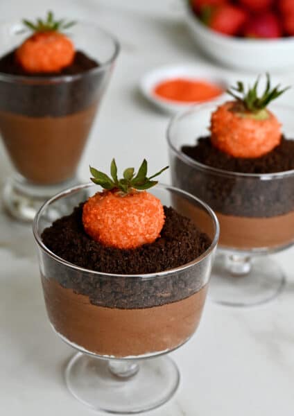 Three chocolate mousse parfaits topped with crushed chocolate wafers and strawberries dipped in orange candy melts. A bowl of strawberries sit behind the parfaits.