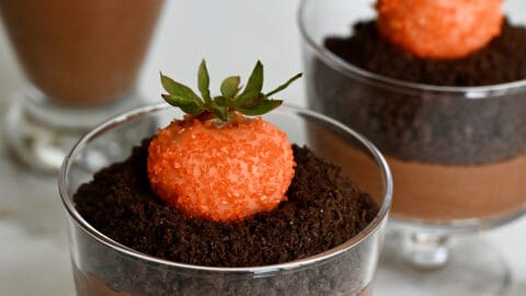 Three chocolate mousse parfaits topped with crushed chocolate wafers and strawberries dipped in orange candy melts. A bowl of strawberries sit behind the parfaits.
