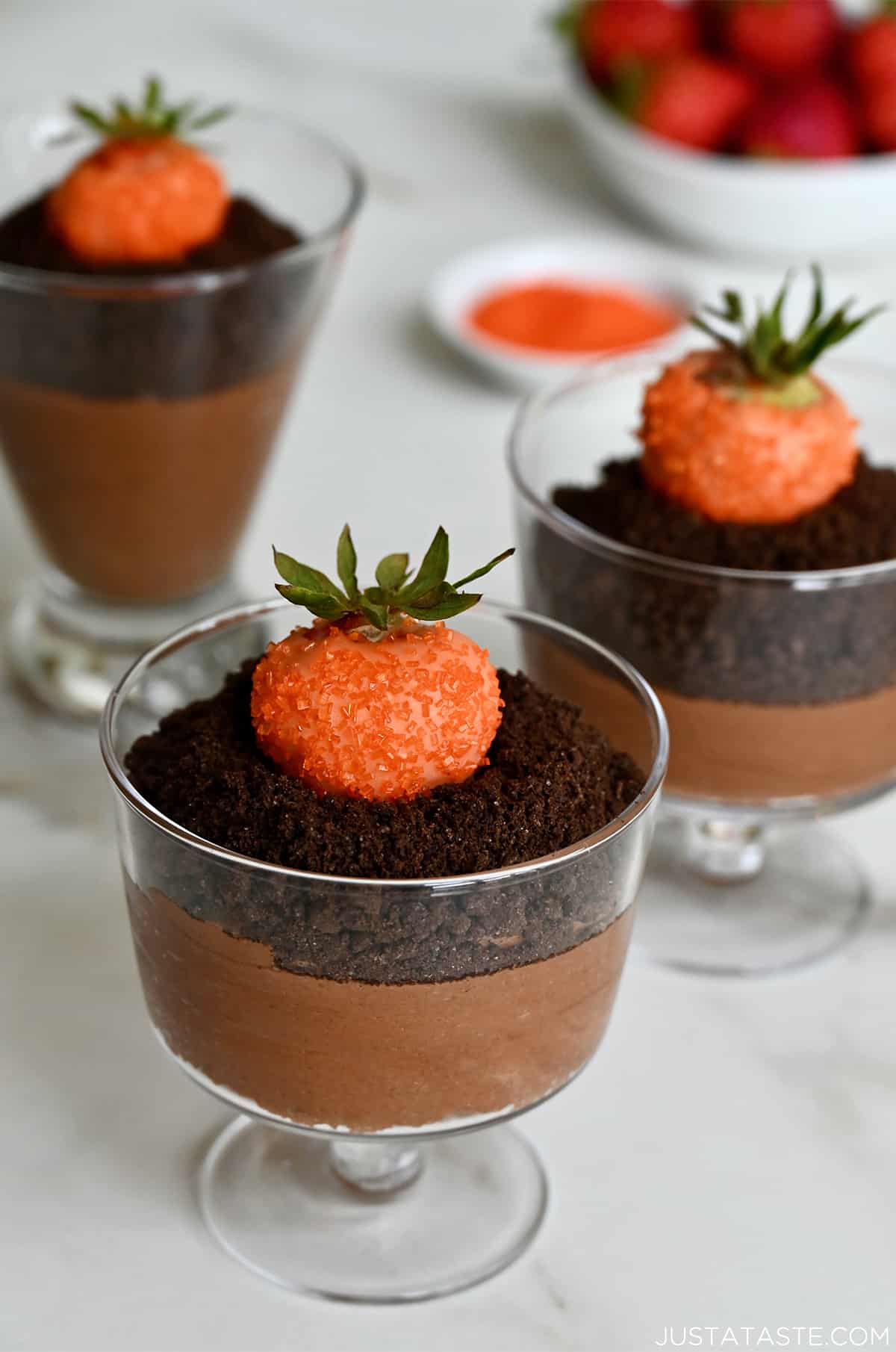 Three chocolate mousse parfaits topped with crushed chocolate wafers and strawberries dipped in orange candy melts. A bowl of strawberries sit behind the parfaits.
