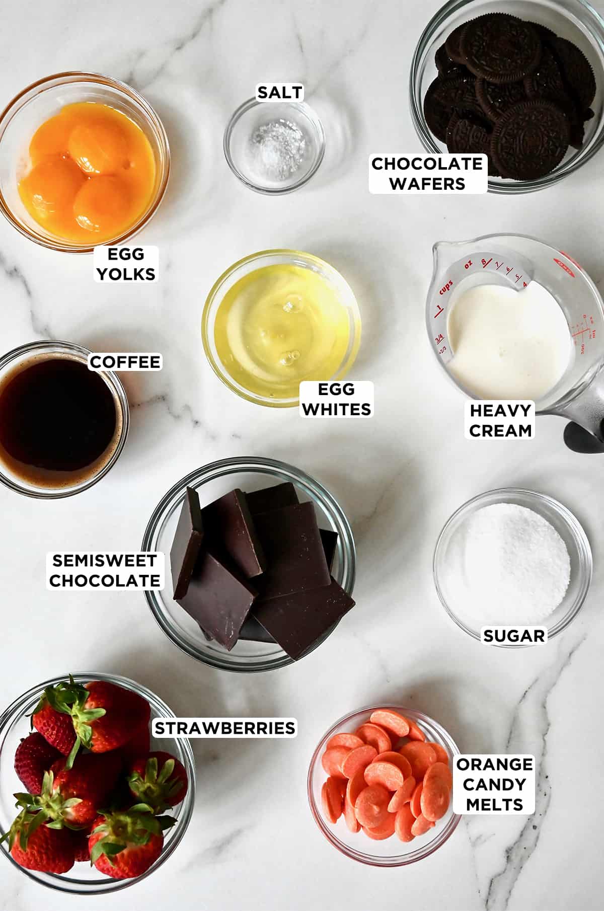 Small glass bowls of strawberries, orange candy melts, sugar, chocolate, coffee, egg whites, heavy cream, chocolate wafers, egg whites and yolks, and salt.