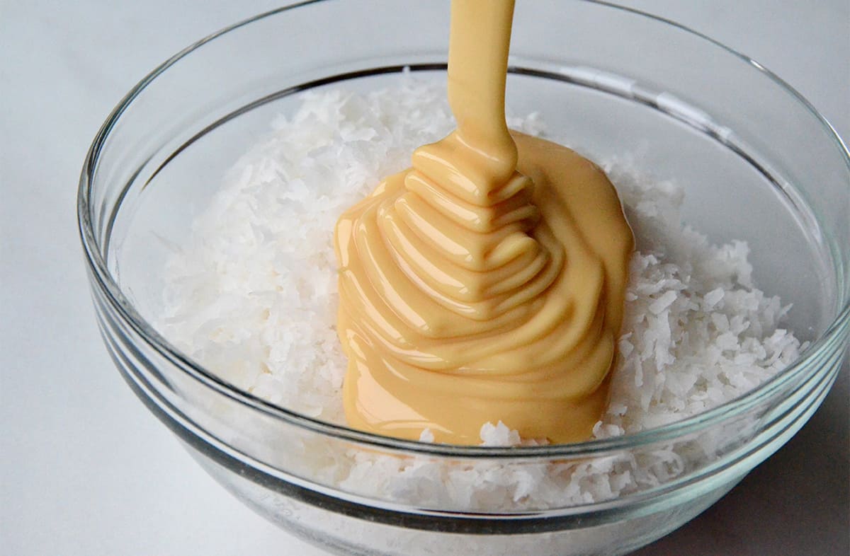 Sweetened condensed milk is drizzled over coconut in a glass bowl.