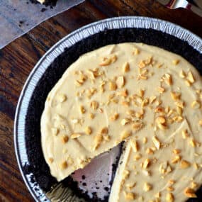 A frozen peanut butter pie topped with chopped peanuts. A slice is taken out of the pie.