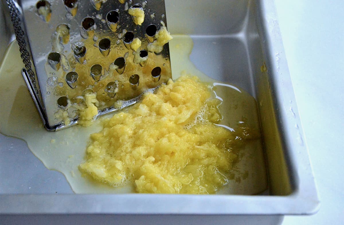 Fresh pineapple is being grated with a box grater into a metal baking dish.