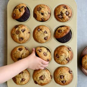 A child's hand reaches for a chocolate chunk Greek yogurt banana muffin in a muffin tin.