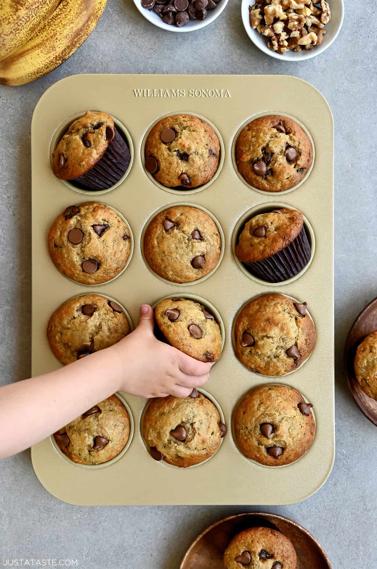 A child's hand reaches for a chocolate chunk Greek yogurt banana muffin in a muffin tin.