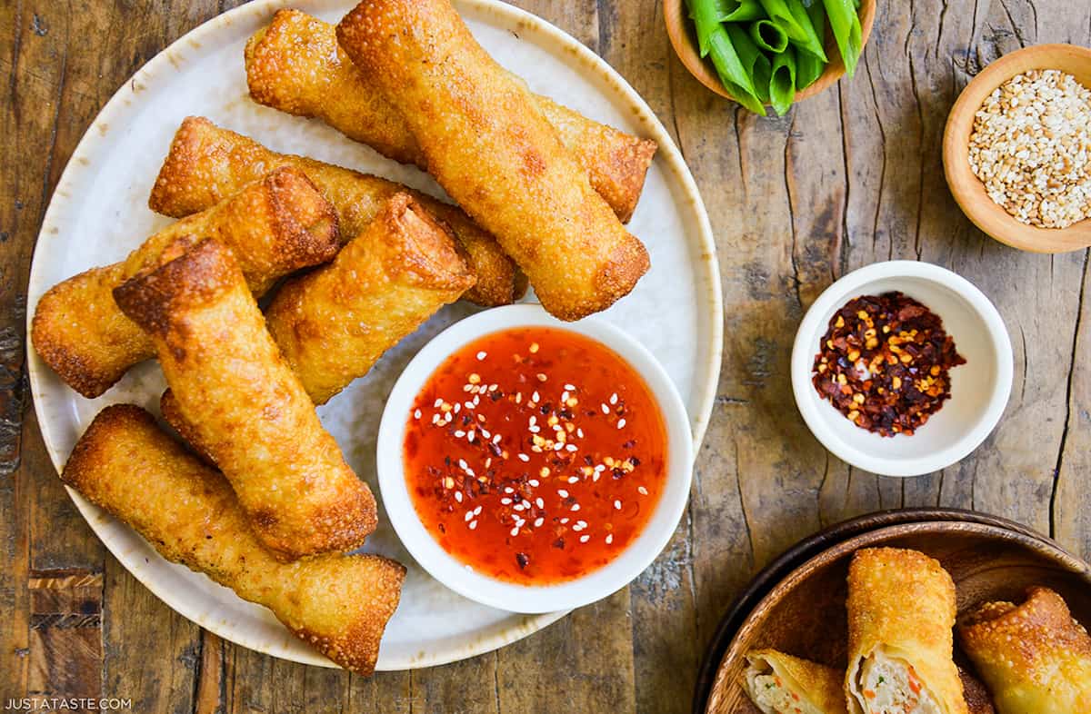 Crispy sesame chicken egg rolls on a plate alongside a small bowl of sweet chili dipping sauce. Small bowls of sliced scallions, sesame seeds and chili flakes are beside the plate.