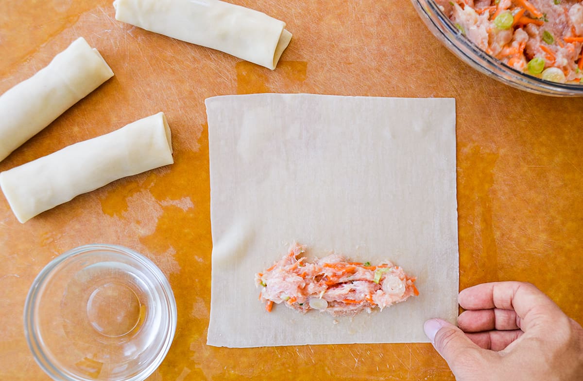 A ground chicken-vegetable mixture on the lower-third of an egg roll wrapper. Rolled egg rolls and a glass bowl with filling are off to the side.