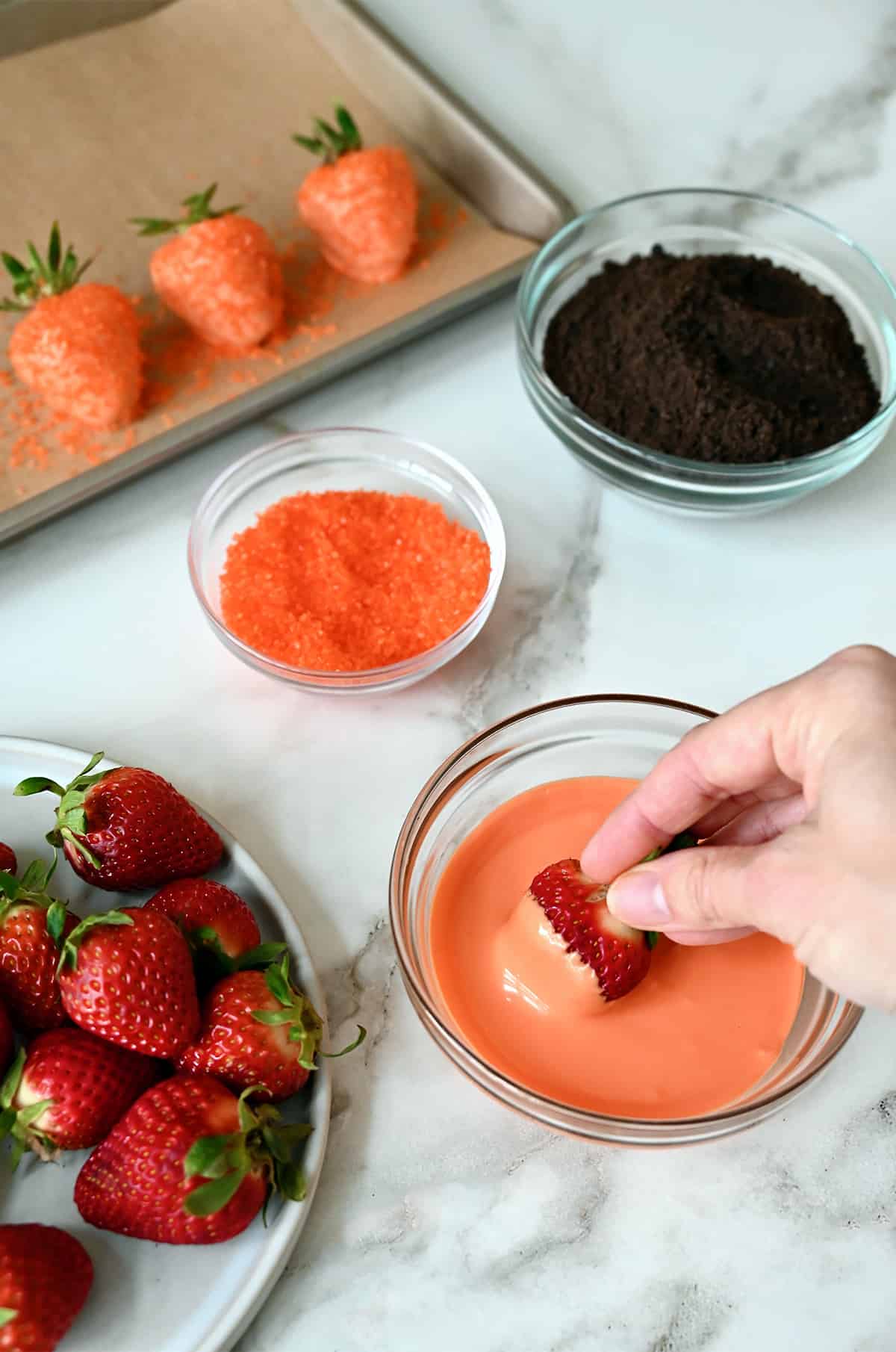 A person dipping a whole strawberry into melted orange candy melts. A plate of whole strawberries and bowls of orange sanding sugar and crushed chocolate wafers are scattered around.