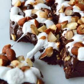 Rock road brownies topped with chopped almonds. A piece of brownie is pulled off the corner.