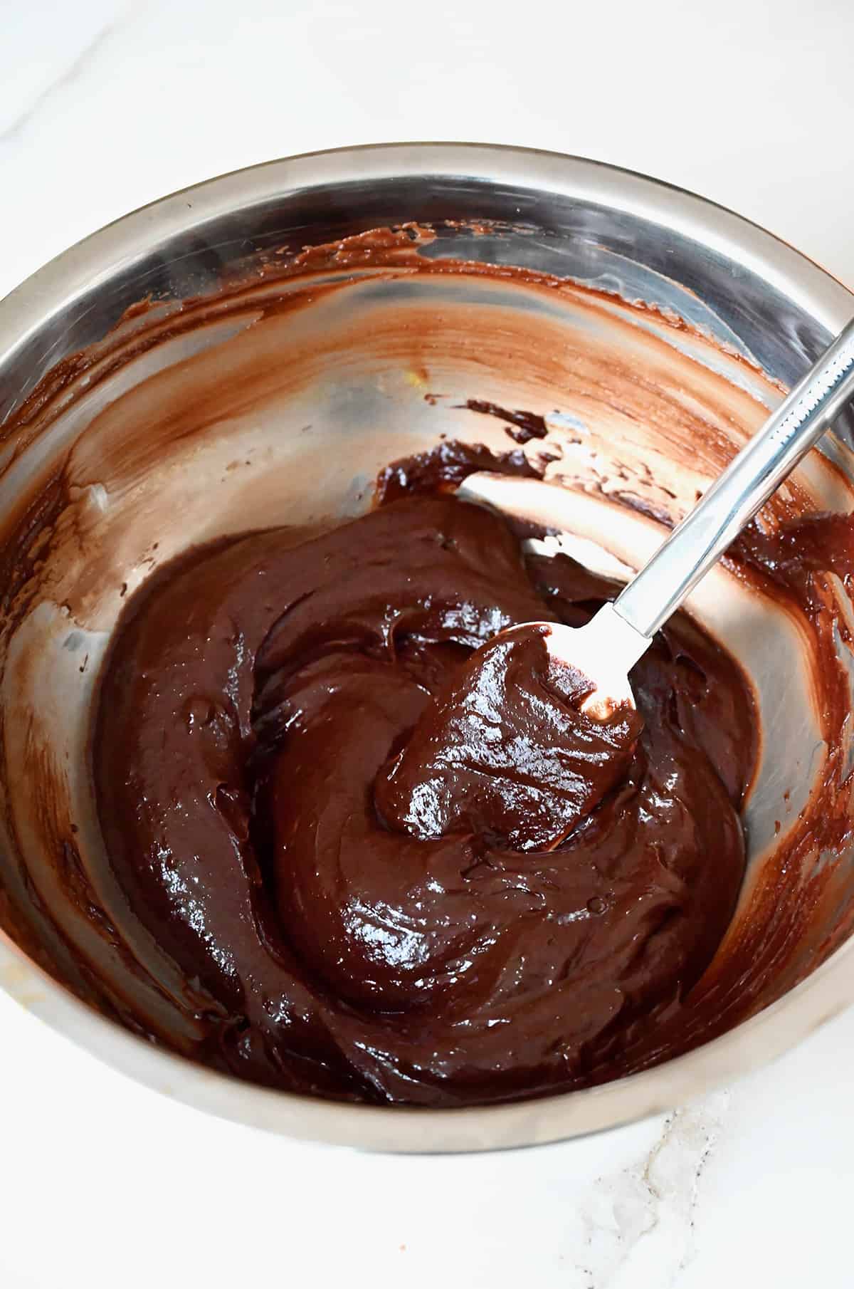 The melted chocolate mixture in a stainless steel mixing bowl with a rubber resting against its side.
