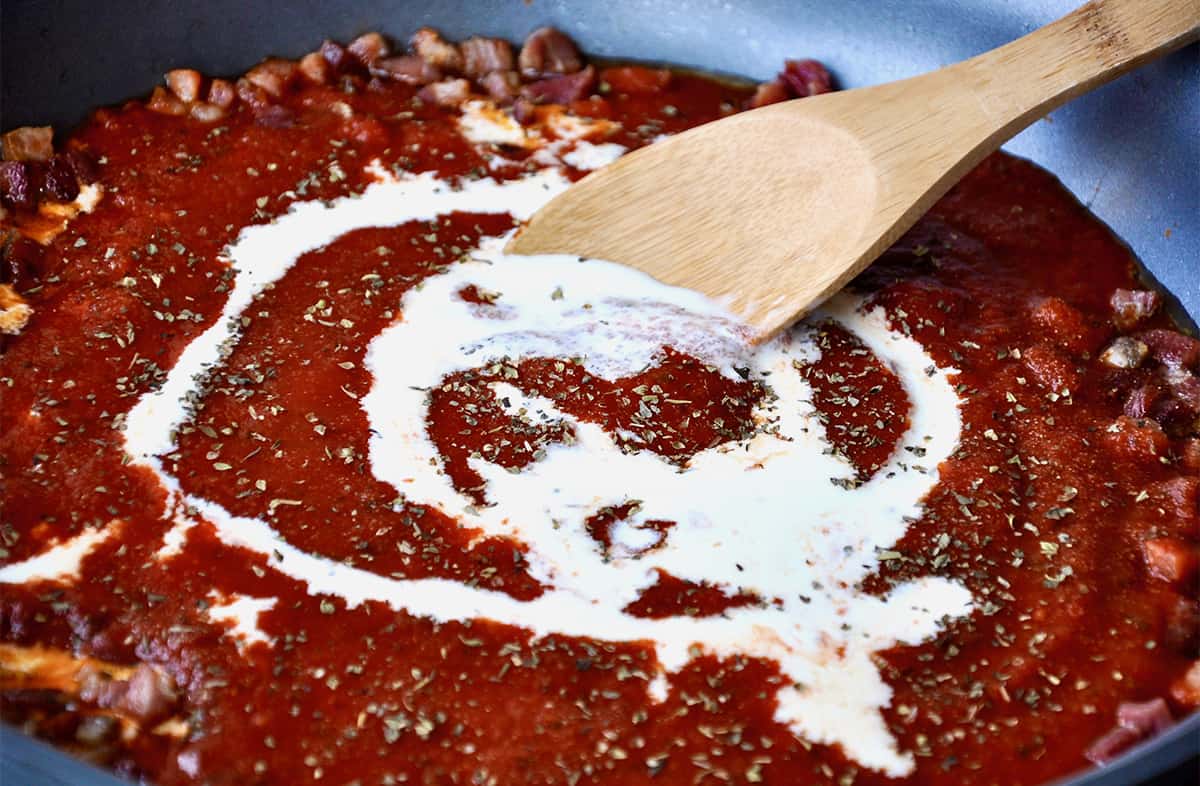 Heavy cream is being stirred into a pan of marinara sauce with crispy pancetta.