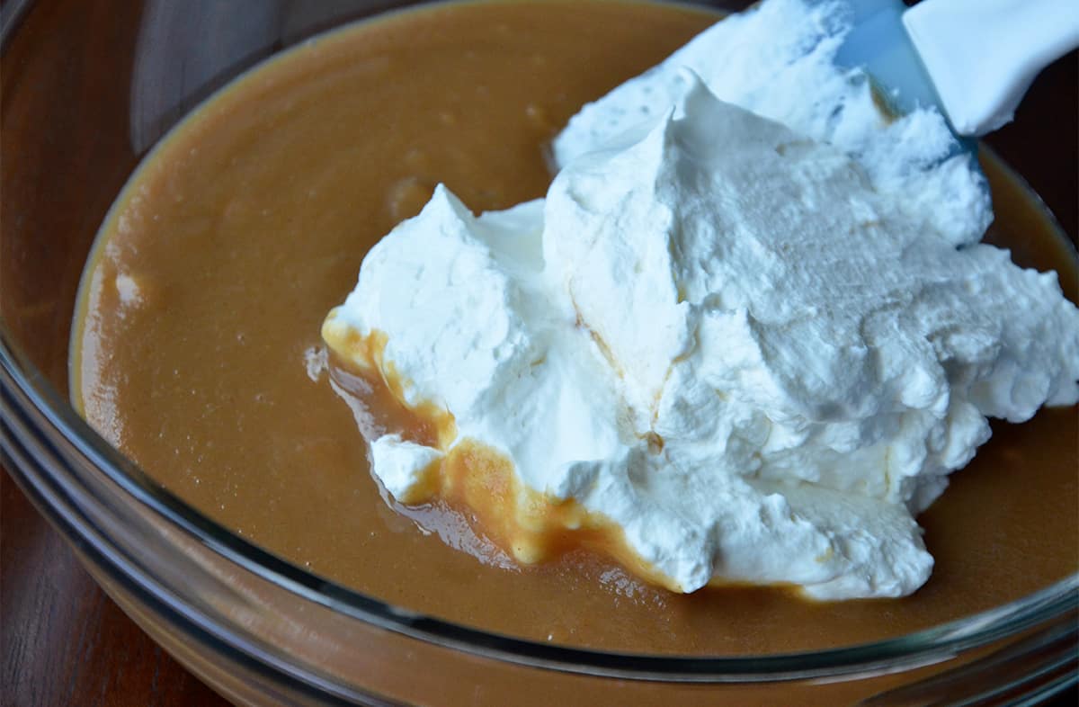 Whipped cream in a peanut butter mixture in a glass mixing bowl. A rubber spatula is propped up against the side of the bowl.