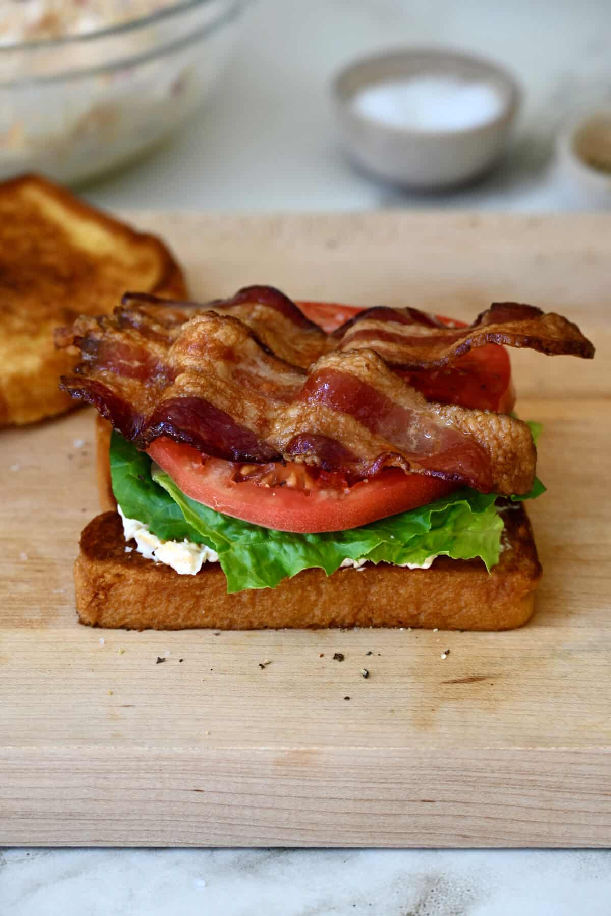 A slice of bread topped with pimento cheese, romaine lettuce, slices of tomato and two crispy bacon slices on a wood cutting board beside a slice of toast.