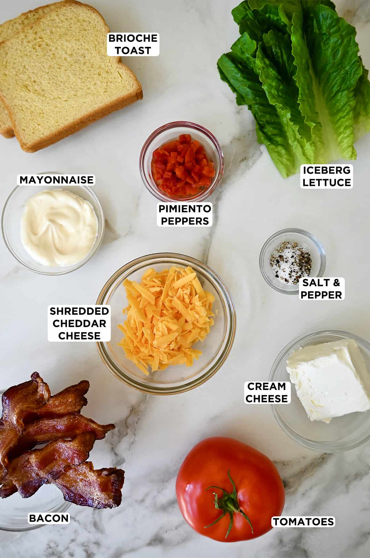 Pimento cheese BLT ingredients in various sizes of glass bowls, including mayo, pimento peppers, salt and pepper, shredded cheddar cheese, cream cheese, romaine lettuce, crispy bacon, tomato slices and slices of brioche bread.