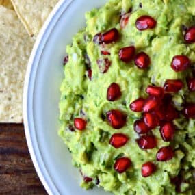 A bowl of pomegranate guacamole topped with extra pomegranate seeds. Tortilla chips are beside the bowl.