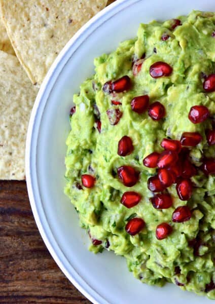 A bowl of pomegranate guacamole topped with extra pomegranate seeds. Tortilla chips are beside the bowl.