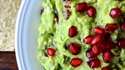 A bowl of pomegranate guacamole topped with extra pomegranate seeds. Tortilla chips are beside the bowl.