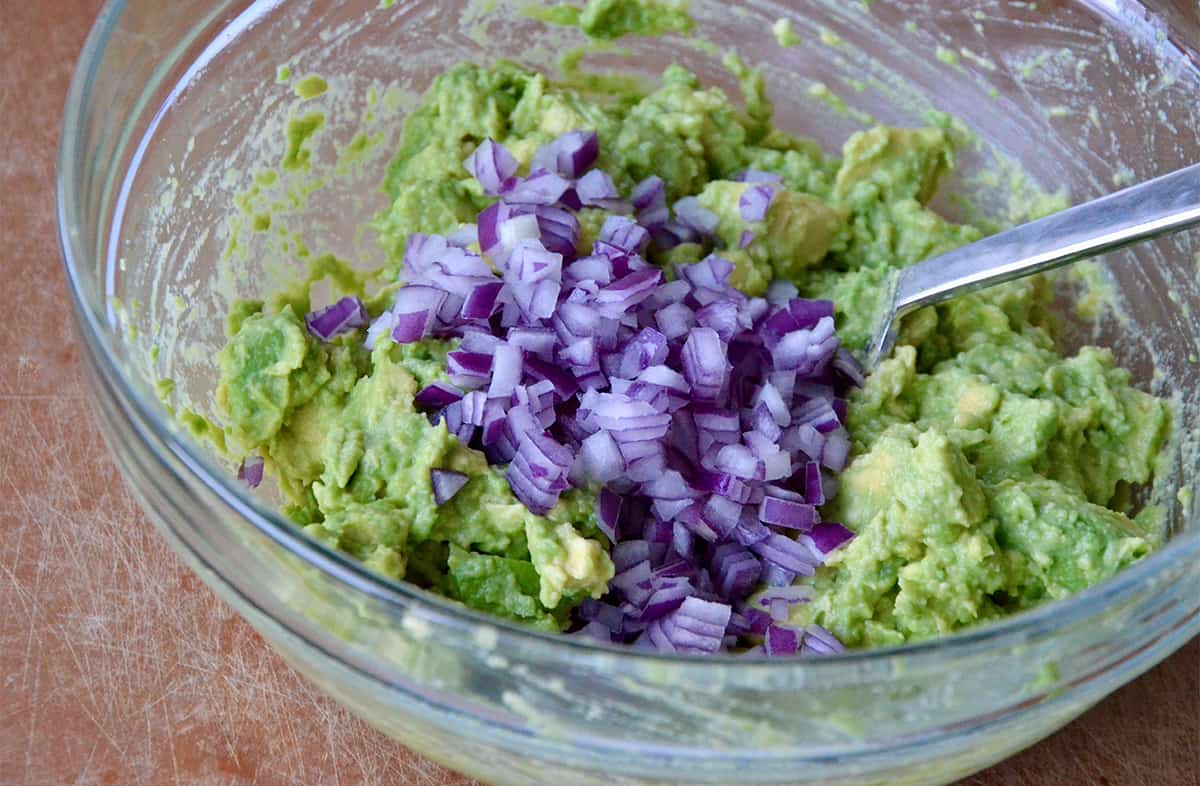 Diced red onion on top of mashed avocado in a glass mixing bowl.