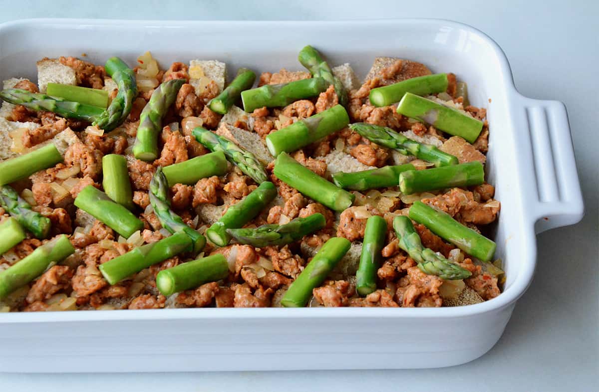An unbaked sausage and asparagus strata in a white casserole dish.