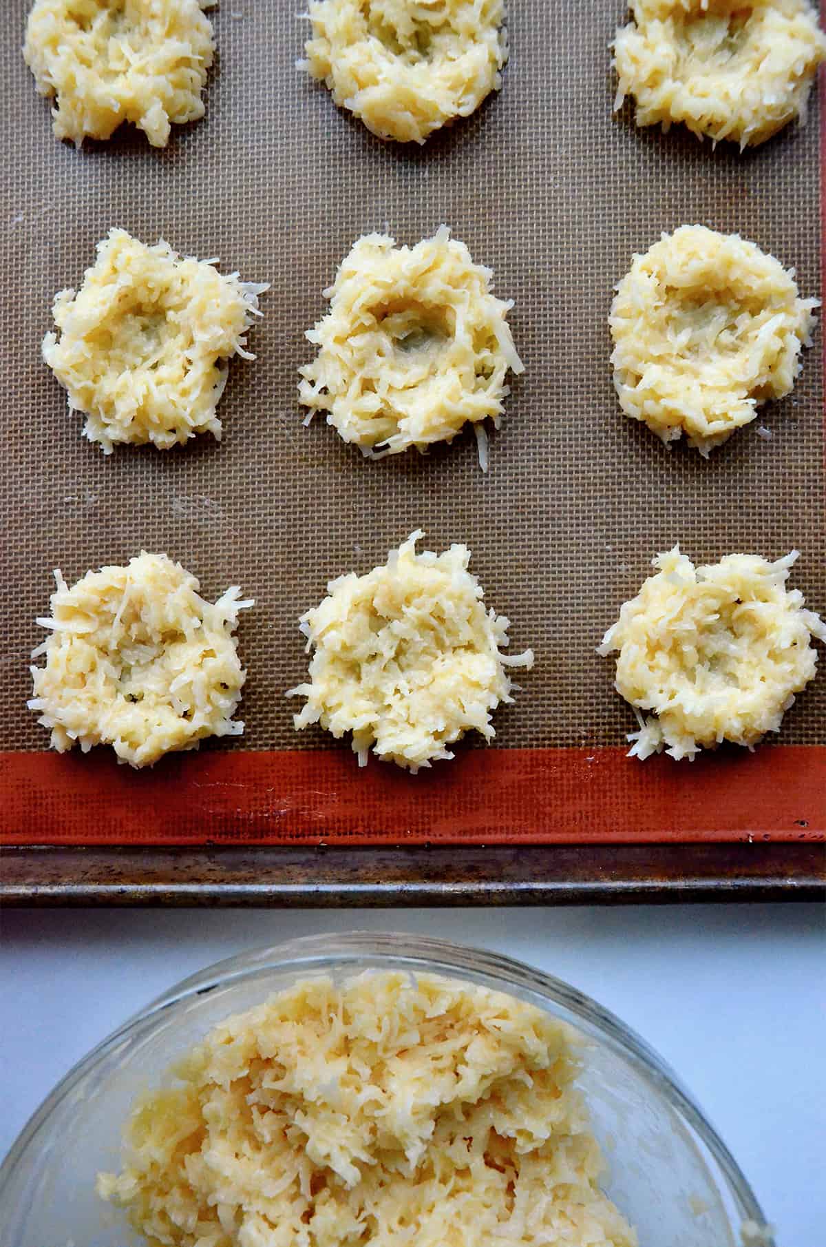 Coconut macaroon nests are lined up on a silpat baking sheet, ready to be baked. A bowl of macaroon batter in a glass bowl is nearby.