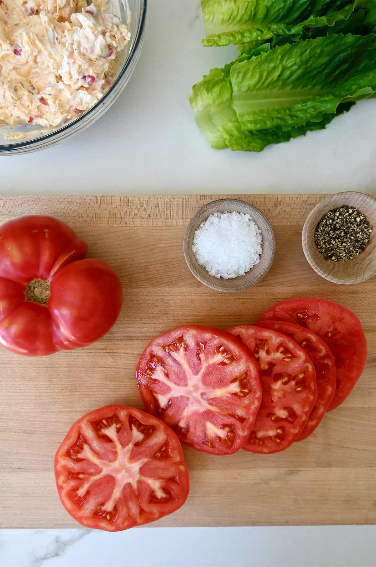 Five slices of tomato next to a whole beefsteak tomato and small bowls containing salt and pepper on a wood cutting board.