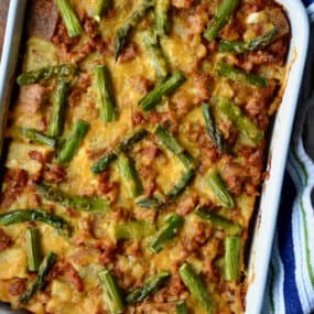 A casserole dish holding a baked breakfast strata with sausage and asparagus. The dish sits on a kitchen towel on a wooden table.