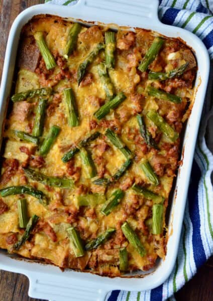A casserole dish holding a baked breakfast strata with sausage and asparagus. The dish sits on a kitchen towel on a wooden table.