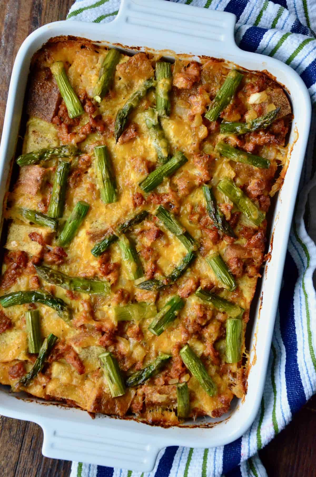 A casserole dish holding a baked breakfast strata with sausage and asparagus. The dish sits on a kitchen towel on a wooden table.