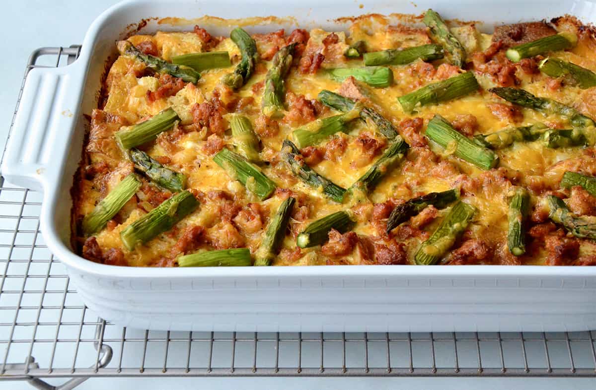 A casserole dish on a wire cooling rack holding a baked breakfast strata with sausage and asparagus.