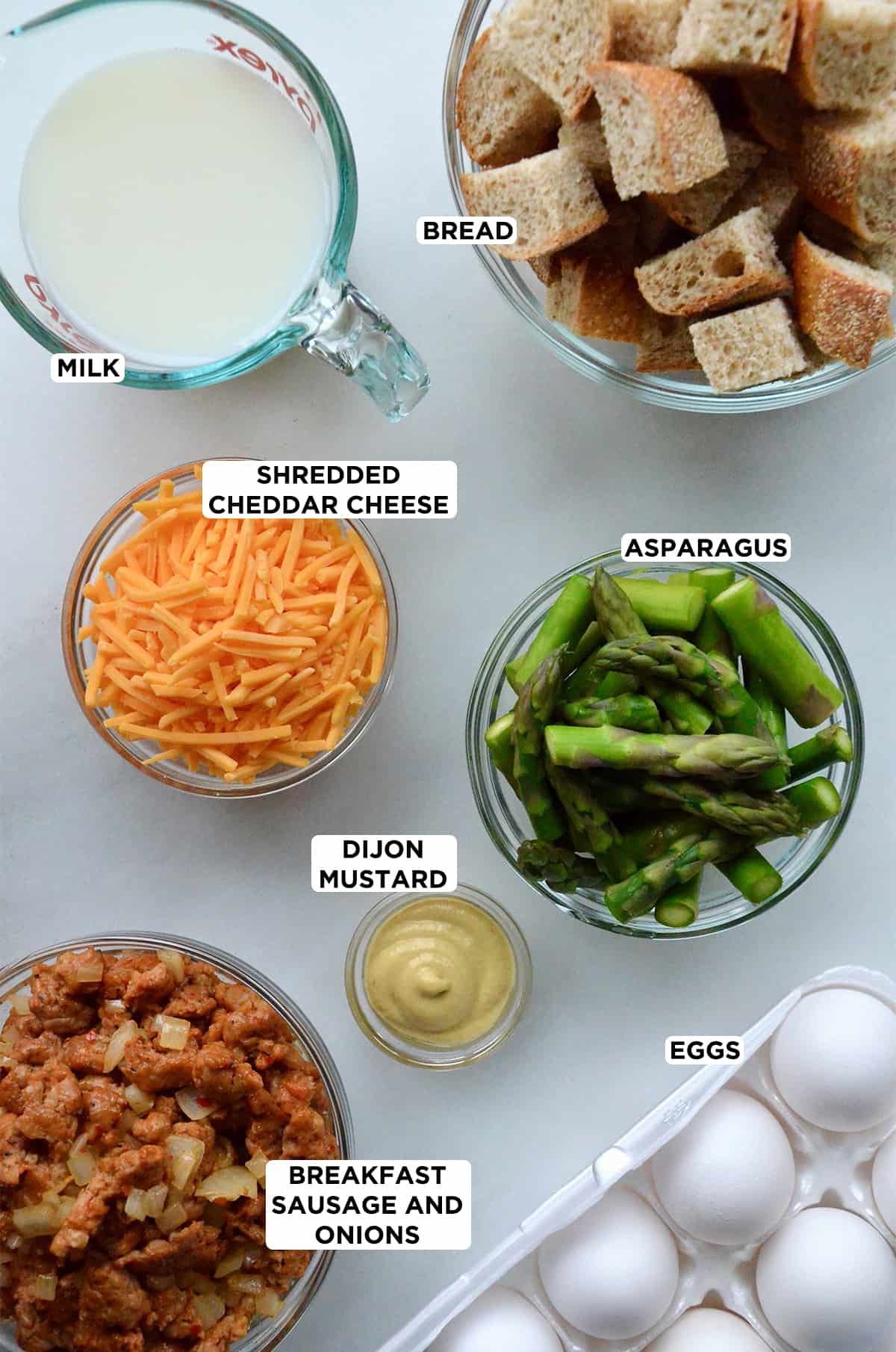 A carton of eggs, a measuring cup of milk and glass bowls of sautéed breakfast sausage and onions, mustard, asparagus pieces, grated cheddar cheese and cubes of bread.