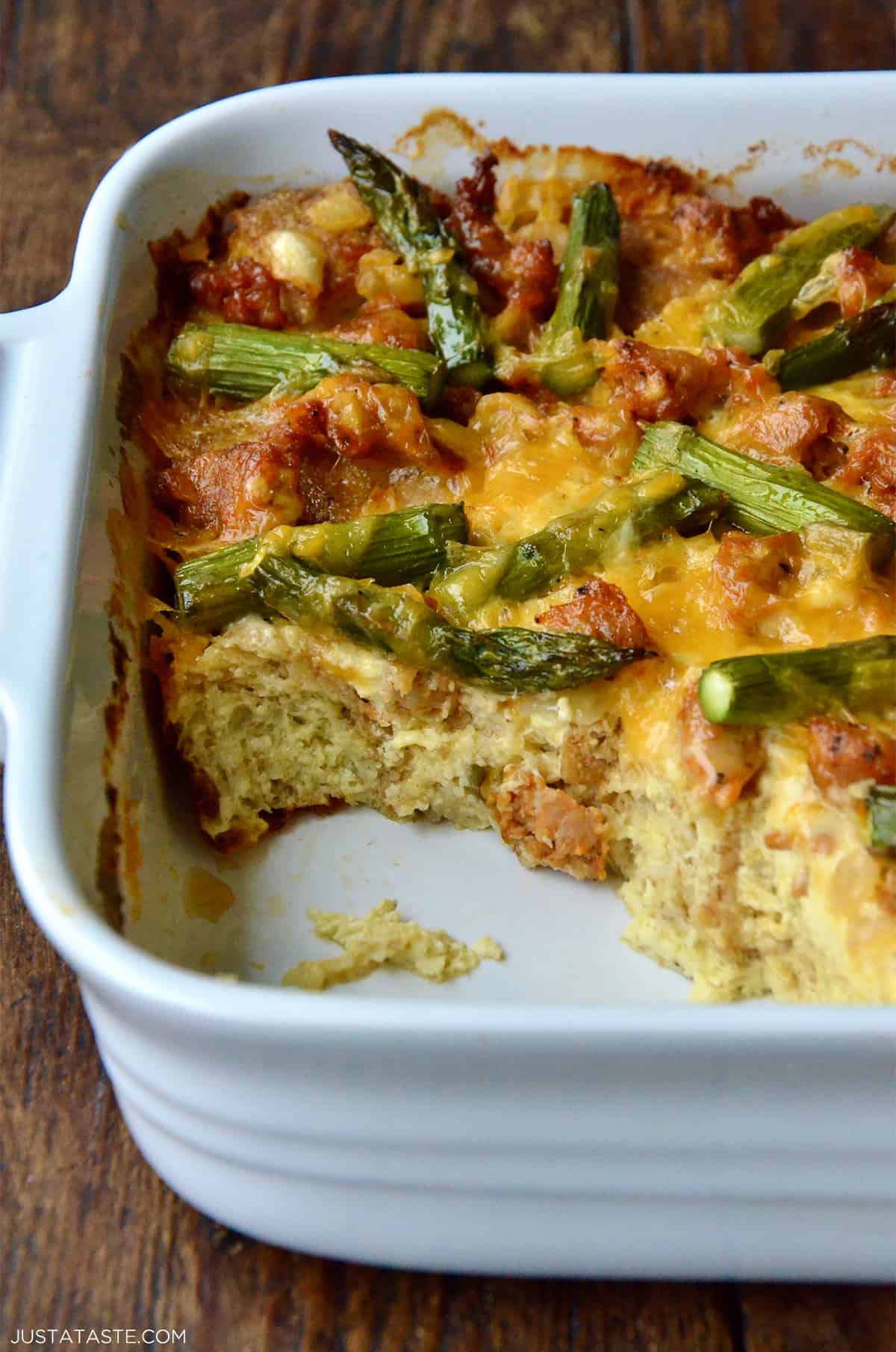 A casserole dish with a baked strata with sausage and asparagus with a large scoop taken out of it.