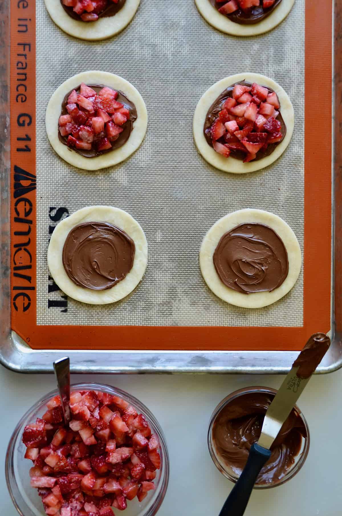 Rounds of pie dough are being filled with Nutella and strawberries. Small glass bowls with diced strawberries and Nutella sit nearby.